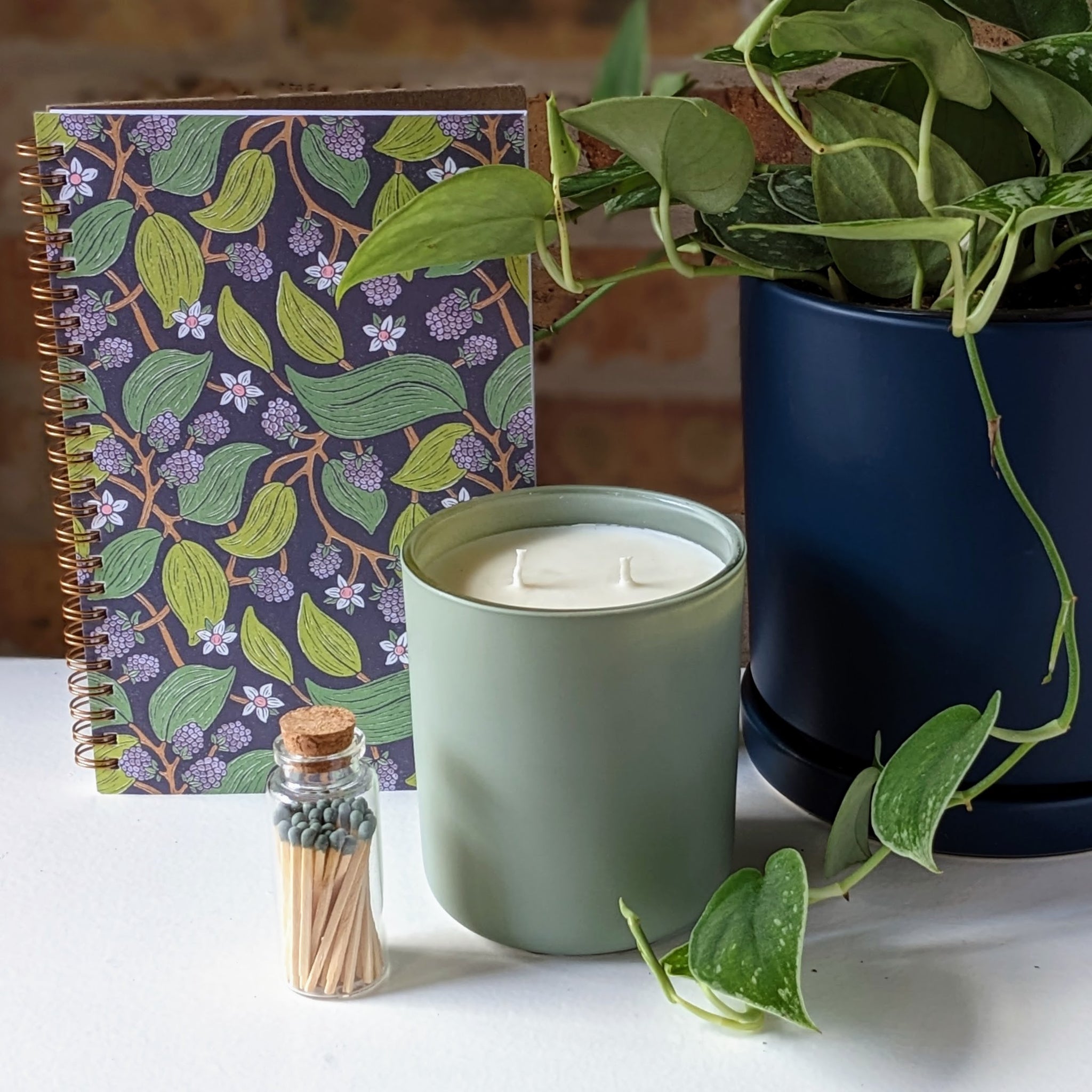 Image of notebook with flowers and leaves design, candle with sage green container, matches in a small glass bottle and pothos in a dark pot for a slow living scene